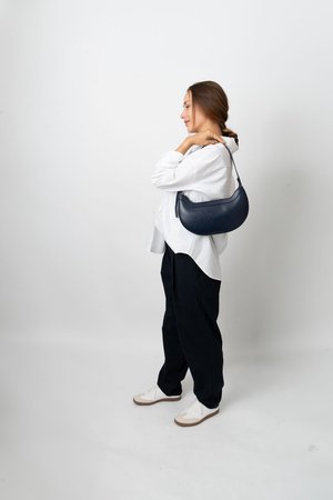 Kvinde iført hvid skjorte og mørke bukser bærer en mørkeblå håndtaske over skulderen og står foran en ensfarvet hvid baggrund.