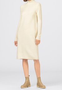 Robe-pull en maille crème avec manches longues et col roulé côtelé. Longueur au genou, coupe décontractée et design minimaliste. Portée avec des bottines beige.