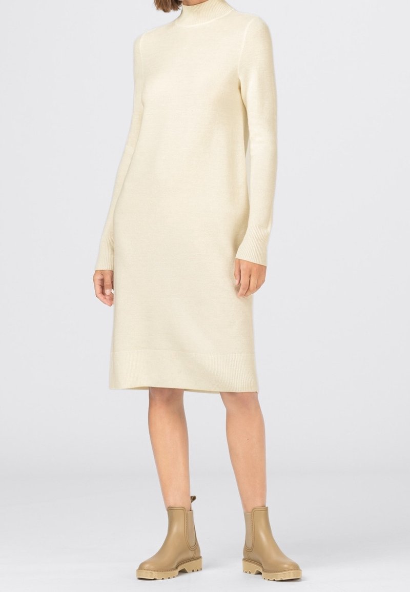 Robe-pull en maille crème avec manches longues et col roulé côtelé. Longueur au genou, coupe décontractée et design minimaliste. Portée avec des bottines beige.