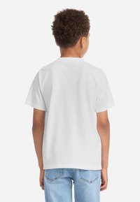 Fehér pamut póló rövid ujjakkal és kerek nyakkal, hátulról nézve. Világoskék farmernadrággal párosítva.