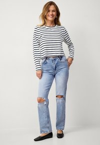 Gestreepte langearmige top in wit en marineblauw, gecombineerd met lichtblauwe ripped jeans. Model draagt zwarte platte schoenen en staat tegen een plain achtergrond.