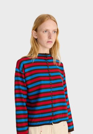 Jeune femme aux longs cheveux roux portant un pull boutonné à rayures horizontales bleu, rouge et marron et un pantalon clair.