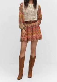 Bunt gemustertes Kleid mit einem cremfarbenen Häkeloberteil, tailliertem Bund und fließendem Rock; kombiniert mit braunen Wildledercowboystiefeln.