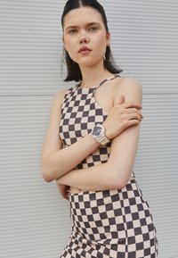 Ensemble crop top et jupe à carreaux en beige et marron, avec un design à col halter. Porté avec une montre claire et de grandes boucles d'oreilles créoles.