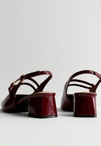 Scarpe slingback a tacco basso in vernice rossa scura con cinturini allacciati, viste da dietro su una superficie bianca semplice.