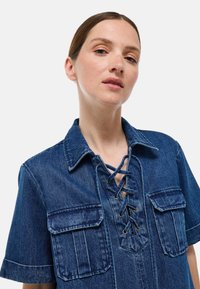 Chemise en denim bleu foncé avec un décolleté lacé, des manches courtes et deux poches poitrine avec des détails de surpiqûres.