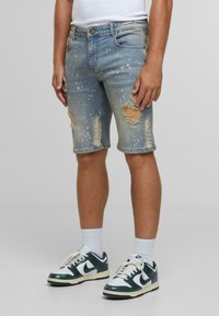 Traper šortsevi svijetloplave boje s istrošenim detaljima, koji uključuju poderotine, prskanje boje i klasičan dizajn s pet džepova.