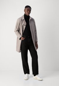 Vaaleanharmaa pitkä trench-coat, jossa on lovettu kaulus, musta poolopaita, mustat housut ja valkoiset lenkkarit, joissa on beiget yksityiskohdat. Suuri pehmeä pinta.