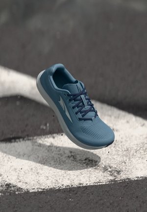 Blauwe sportschoen met donkerblauwe veters zweeft boven asfalt met witte markeringen, kleine stukjes gruis vallen eronder.