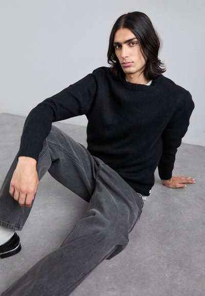 Pulover negru tricotat cu guler rotund și manșete pliate, asortat cu pantaloni din denim gri lejeri și pantofi negri pe o suprafață gri.