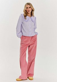 Fioletowa koszula w kratkę z dużym falbaniastym kołnierzem, zestawiona z różowymi szerokimi spodniami z korduroju. Żółte buty wsuwane dopełniają całość.