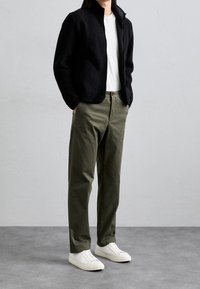 Chaqueta negra con textura y cremallera sobre una camiseta blanca, combinada con pantalones verde oliva y zapatillas blancas. El diseño es moderno y casual.