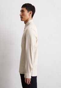 Beige Langarmhemd mit Button-down-Kragen, aus einem glatten Stoff gefertigt, mit geradem Saum und gemanschetten Ärmeln.