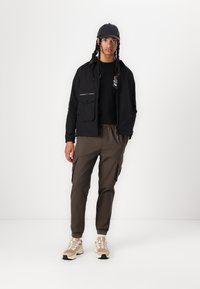 Black lightweight jacket with zip pockets, paired with a black t-shirt and olive green cargo pants. Light beige sneakers complete the look.