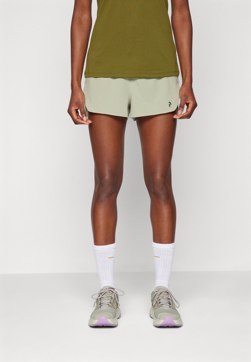 Athlète portant un débardeur vert olive, un short de course vert clair, des chaussettes blanches montantes et des chaussures de course grises à semelles violettes, debout.