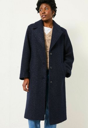 Marineblauer Wollmischungs-Mantel mit strukturiertem Finish, großen Revers, Knopfverschluss und aufgesetzten Taschen, getragen über einem leichten Pullover und Jeans.