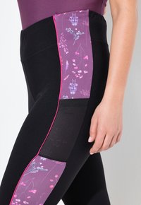 Zwarte leggings met een paarse bloemenpaneel aan de zijkant en roze accenten, gemaakt van rekbare stof. Mesh-textuur inzet op het kuitgedeelte. Gladde tailleband.