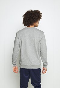 Sudadera gris de cuello redondo con mangas largas, puños y bajo de canalé. Textura suave, diseño liso, llevada con pantalones azul marino.