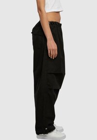Pantalons cargo noirs avec taille élastique à cordon de serrage, poches latérales et poignets élastiques. Associés à des baskets blanches, présentés de profil.