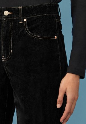 Czarne spodnie sztruksowe o subtelnej teksturze, z miedzianymi akcentami, widocznymi stębnowaniami i przednimi kieszeniami, prezentowane na niebieskim tle.