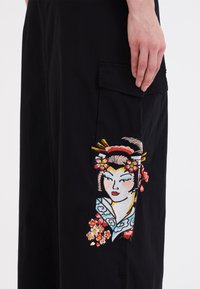 Sorte cargo bukser med farverigt broderet design af en kvinde med blomster og hårnåle, tekstureret stof og sidelommer.