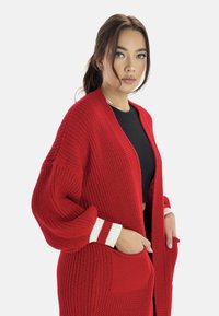Cardigan de punto rojo con mangas oversized, acentos de rayas blancas en los puños y bolsillos. Tejido texturizado con un diseño de frente abierto.