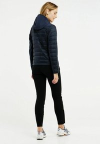 Námornícka prešívaná bunda s kapucňou, čierne legíny. Modelka zobrazuje spätný pohľad s tonálnymi akcentmi a športovými topánkami.