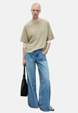 Beige oversized t-shirt with short sleeves, paired with wide-leg light blue jeans. Black textured bag, standing on a plain background.