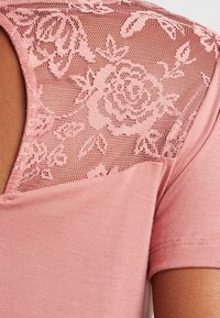 Pink T-shirt med en blondepanel på skuldrene med blomster mønstre. Tøjet er blødt med en glat tekstur og korte ærmer.