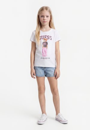 Ragazza giovane con lunghi capelli biondi che indossa una t-shirt grafica bianca, pantaloncini di jeans e scarpe da ginnastica bianche, in piedi su uno sfondo bianco.