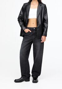 Person trägt eine schwarze Lederblazer, ein weißes kurz geschnittenes Oberteil, lose dunkle graue Jeans und schwarze Stiefeletten, steht mit einer Hand in der Tasche.