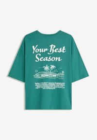Grøn bomulds-T-shirt med korte ærmer. Bagsiden har hvid tekst og en illustration af et hus med palmer, der fremhæver et strandtema.