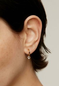 Pendiente de aro dorado con una gema redonda clara. El pendiente presenta un acabado suave y un diseño sencillo, colgando cerca de la oreja.