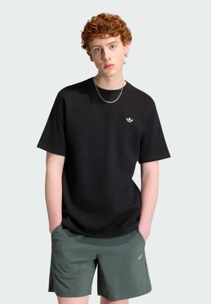 Giovane uomo con capelli rossi e ricci che indossa una maglietta nera Adidas e pantaloncini verdi Adidas, in piedi con le mani nelle tasche davanti a uno sfondo semplice.