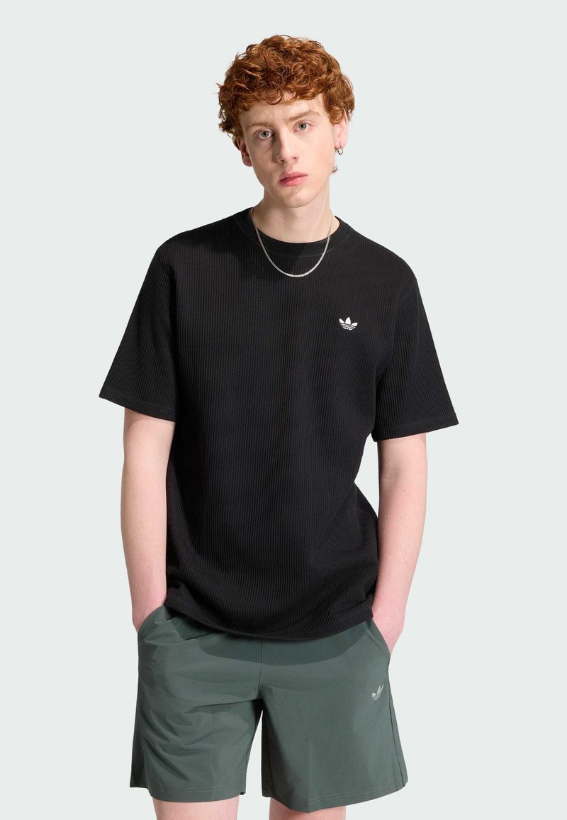 Jeune homme aux cheveux roux bouclés portant un t-shirt noir Adidas et un short vert Adidas, debout les mains dans les poches devant un fond uni.