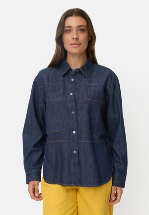 Kvinde iført en mørkeblå denim skjorte med knapper og lange ærmer samt gule bukser, stående mod en ensfarvet lys baggrund.