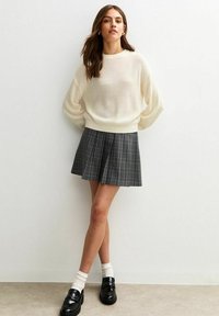 Maglione rigato beige chiaro con spalle cadenti, abbinato a una gonna plissettata a quadri neri e mocassini neri, accessoriato con calzini bianchi.