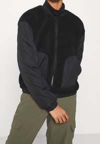 Blouson aviateur noir avec un corps duveteux, des manches matelassées et une fermeture éclair. Il comprend des accents en jersey et des poches latérales.