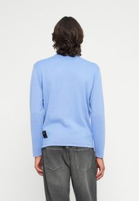 Suéter de mangas longas azul claro em tecido suave. Apresenta um decote redondo e um rótulo de marca discreto no lado inferior esquerdo. Usado com jeans cinzas.