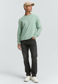 Lichtgroene sweatshirt met ronde hals en bedrukte tekst, gecombineerd met donkere jeans en witte sneakers. Vergezeld van een beige honkbalpet.