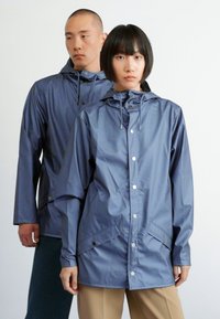 Vestes imperméables bleu marine avec capuchons, boutons-pression et poches avant, portées par deux personnes sur un fond uni.