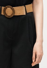 Personne portant un pantalon noir et une ceinture tissée beige avec une grande boucle ronde en bois, bras détendu le long du corps.