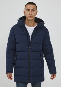 Marineblauer Pufferjacke mit Kapuze, ausgestattet mit einem Reißverschluss, zwei Seitentaschen und horizontalen Steppnähten für zusätzliche Isolierung und Textur.