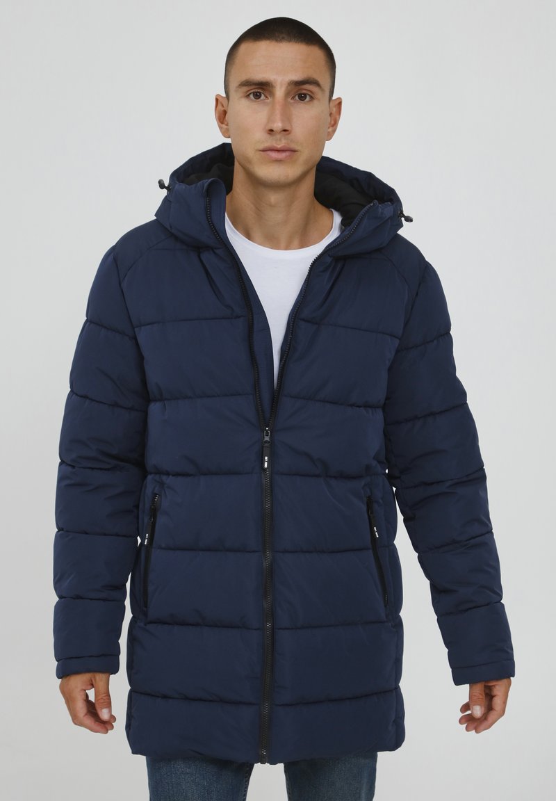 Marineblauer Pufferjacke mit Kapuze, ausgestattet mit einem Reißverschluss, zwei Seitentaschen und horizontalen Steppnähten für zusätzliche Isolierung und Textur.