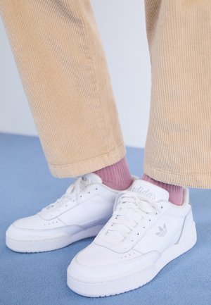 Voeten met witte Adidas-sneakers, lichtpaarse sokken en beige corduroy broeken staand op een blauwe vloer.