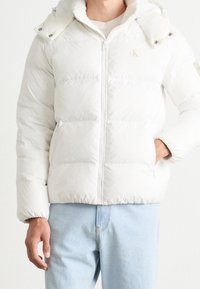 Mężczyzna w białej ocieplanej kurtce z kieszeniami i jasnoniebieskich dżinsach, stoi na tle białego tła.