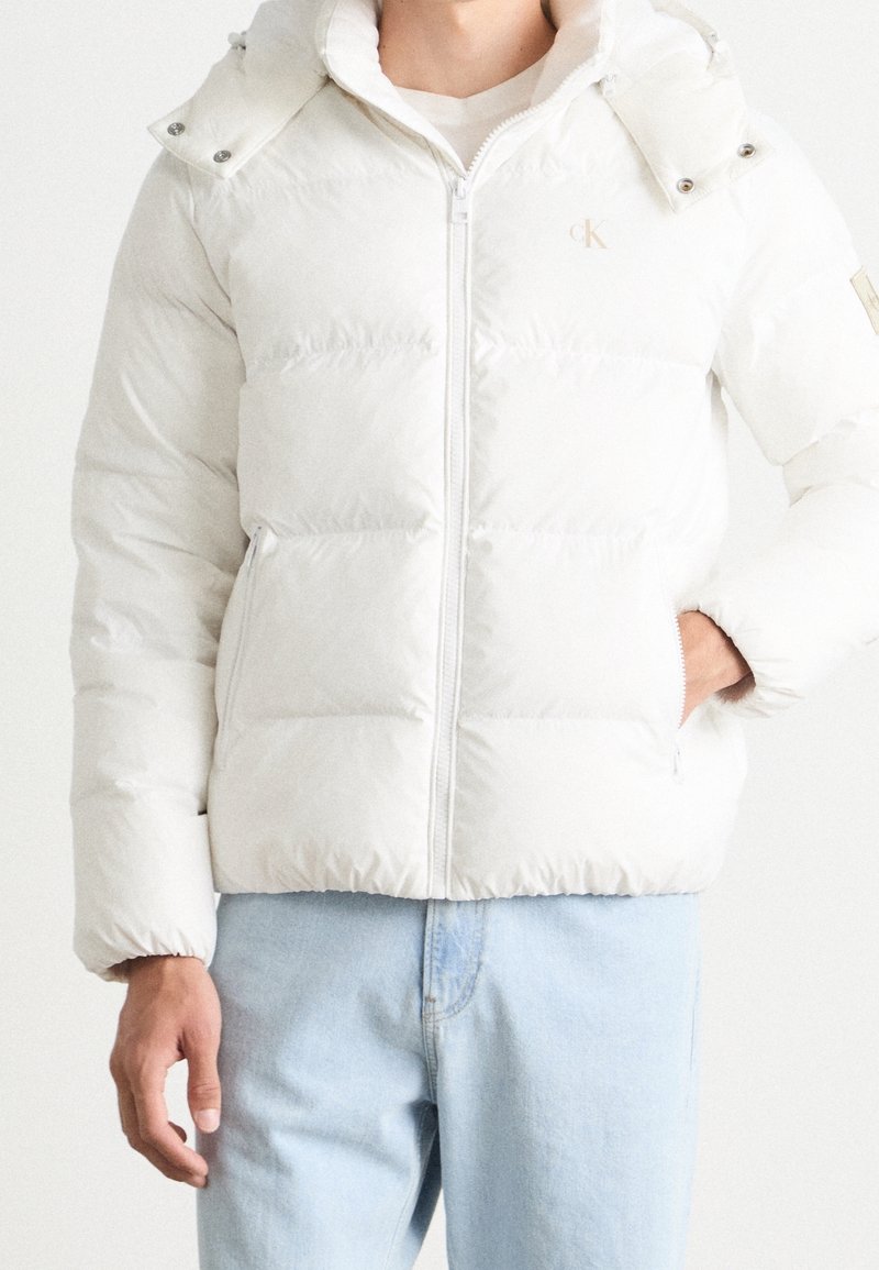 Mężczyzna w białej ocieplanej kurtce z kieszeniami i jasnoniebieskich dżinsach, stoi na tle białego tła.