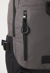 Sac à dos gris avec un motif à carreaux, équipé d'une fermeture éclair et d'un tirant noirs, d'une boucle latérale et d'un tissu texturé.