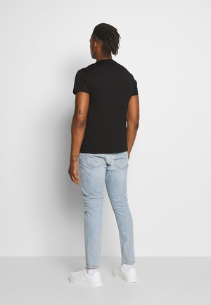 Uomo con dreadlocks che indossa una maglietta nera, jeans azzurri e scarpe da ginnastica bianche, in piedi di fronte a un muro bianco.