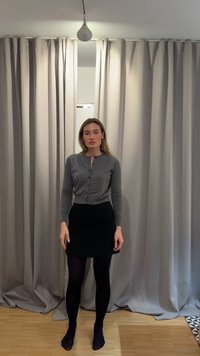 Mujer de pie descalza sobre un suelo de madera, vistiendo un cárdigan gris abotonado, falda negra y medias negras frente a unas cortinas beige cerradas.